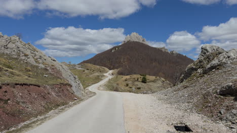 a winding, narrow mountain road carves its path toward the summit, weaving through a rugged landscape beneath a blue, cloud-strewn sky. captured in a wide shot and slow motion on a clear, sunlit day