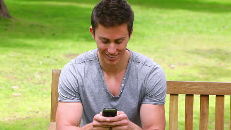 man sits on a bench while looking at at his mobile phone before laughing and looking at camera