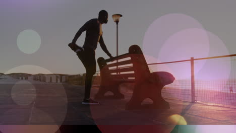 puntos de luz contra el hombre afroamericano en forma estirando la pierna cerca de un banco en el paseo marítimo