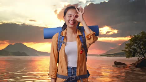 excursionista asiática con mochila de montañismo sonriendo y mostrando el signo ok con los dedos sobre los ojos mientras está de pie en un lago durante la hora del atardecer