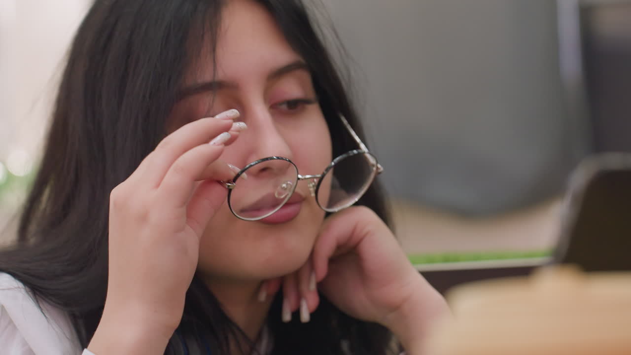 Close up of elegant young woman with long dark hair gently removing glasses while gazing at tablet screen with relaxed, subtle expression, hand supporting her chin, soft indoor background