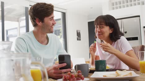 video de una feliz pareja diversa usando un teléfono inteligente y bebiendo café juntos en la cocina