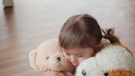 adorable blonde little girl hugs two soft toy teddy bears tight clasp to cheeks and shows toothy smile