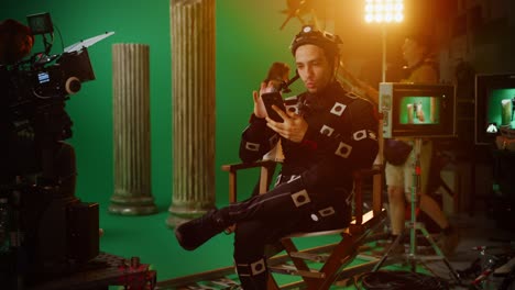 hombre guapo y sonriente con traje de captura de movimiento y equipo de cabeza teniendo descanso para almorzar, sentado en una silla, usa un teléfono inteligente. estudio película de alto presupuesto. en el estudio de cine período de traje drama set de película