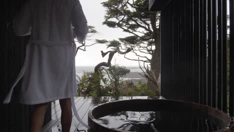 man removes his robe then takes a relaxing bath on wooden hot tub