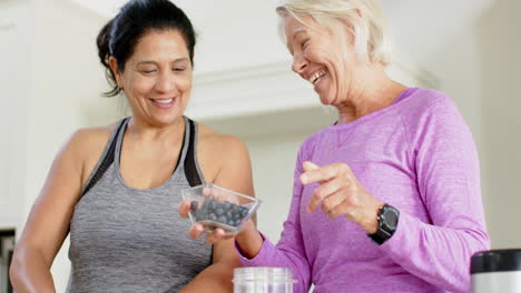 Two-happy-diverse-senior-women-preparing-cocktail,-discussing-and-smiling-in-kitchen,-slow-motion