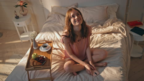 dreamy girl resting bed in morning sunlight. happy teenager holding smartphone