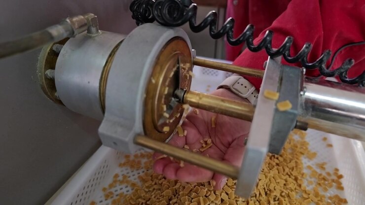 Close-up de uma máquina extrusora de massa despejando pedaços de massa fresca em uma bandeja, enquanto a mão de um trabalhador recolhe o produto durante a produção.