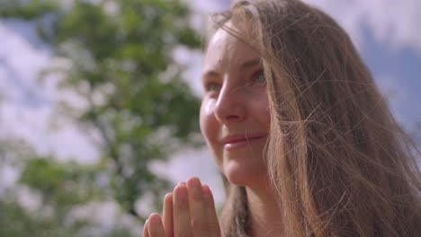 Close-up-of-white-woman,-smiling-with-hands-in-namaste-yoga-pose-in-forest,-green-tree-on-background