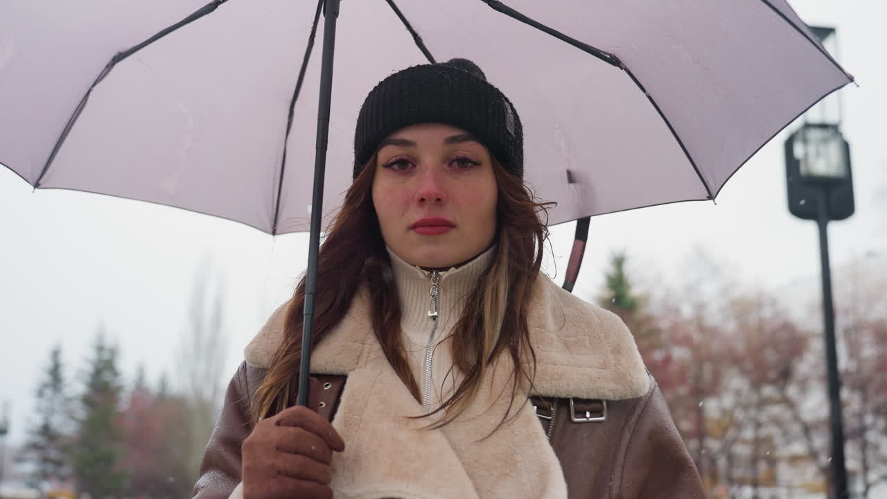 Female with serious expression standing still, holding umbrella, wearing black knit cap and brown shearling jacket, under cold overcast sky with light snowfall, looking contemplative
