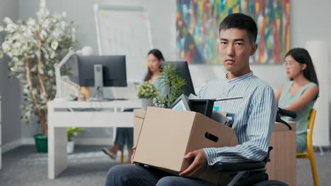 disabled ambitious man in shirt with asian korean beauty sits in wheelchair at desk in the company on knees holding cardboard box with packed things, gets fired from job unable to fulfill his duties