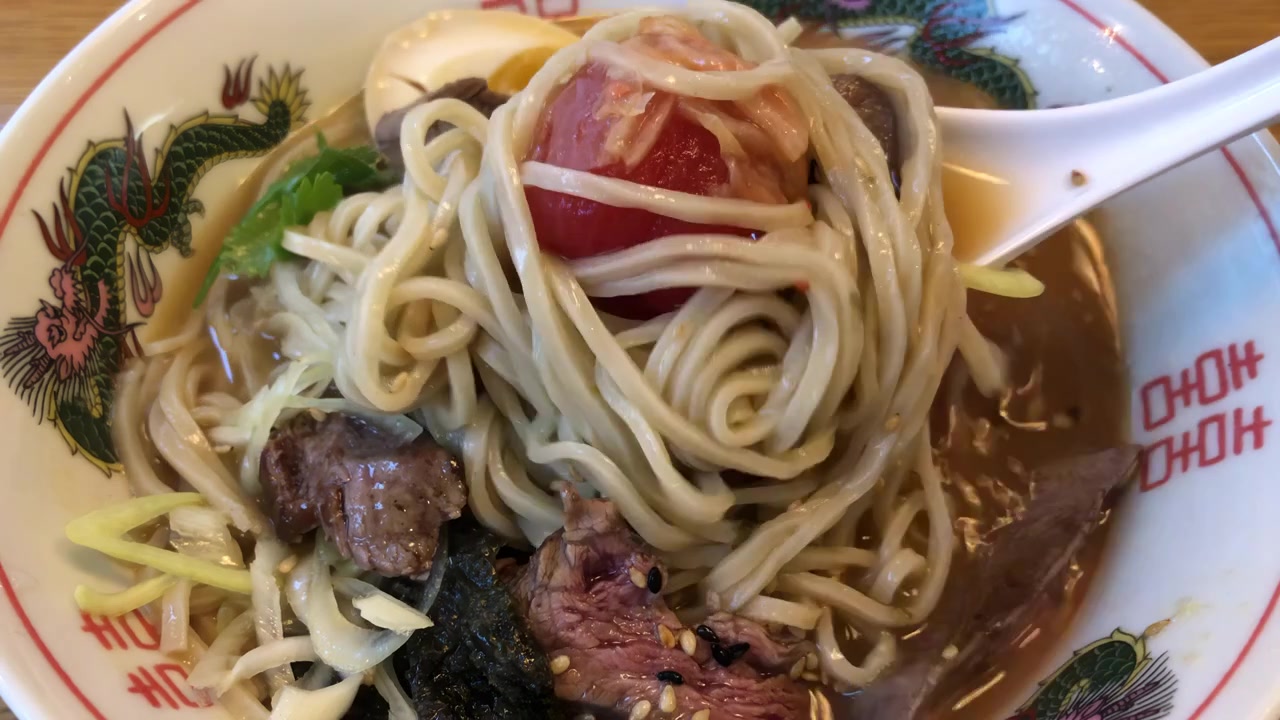 Sendok Jepang mengaduk dalam mangkuk ramen yang berisi irisan daging sapi setengah matang, telur marinasi, tomat ceri, biji wijen, dan herba dalam kuah kaldu yang kaya.