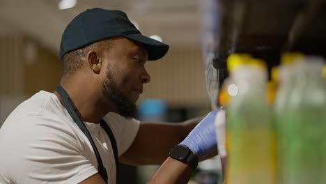 Filmado-De-Lado,-Un-Hombre-De-Piel-Negra-Y-Barba-Con-Una-Gorra-Negra-Y-Una-Camiseta-Blanca-Coloca-Y-Clasifica-Cosas-Y-Botellas-En-El-Mostrador-De-Un-Gran-Supermercado.