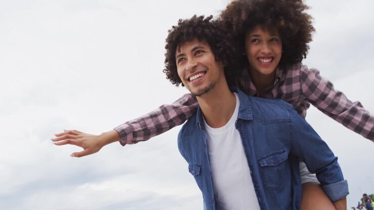 African american man giving his wife a piggyback ride on the promenade near the beach