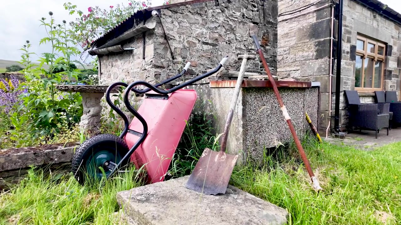 Una carriola arrugginita e delle pale riposano nell'erba alta accanto a una vecchia casa in pietra in un tranquillo cortile rurale.