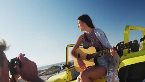 Hombre-Caucásico-Tomando-Fotografías-De-Una-Mujer-Sentada-En-Un-Buggy-De-Playa-Junto-Al-Mar-Tocando-La-Guitarra