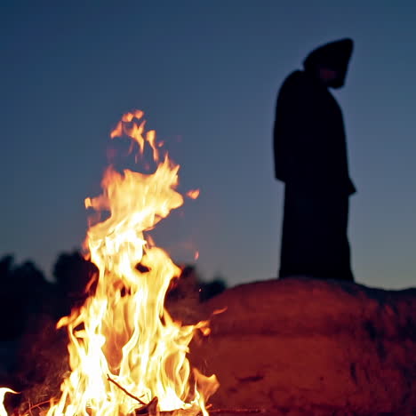 silhouette of black death on the hill at night. flame on the ground and dark figure in witch costume in nature. halloween evening.