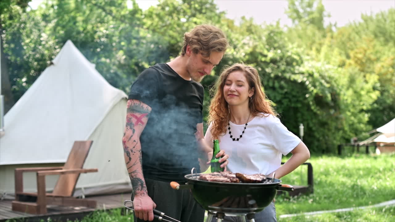 Giovane coppia che frigge carne alla griglia e beve birra, ridendo. Tanto verde intorno. Glamping. Slow motion