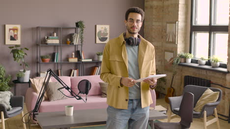 hombre con auriculares de pie sosteniendo papeles en una habitación con micrófono y mirando a la cámara