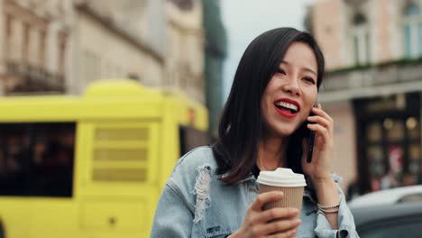 Joven-Alegre-Y-Elegante-Que-Tiene-Una-Agradable-Charla-En-El-Teléfono-Móvil-Mientras-Está-De-Pie-En-El-Centro-De-La-Ciudad-Con-Un-Café-Para-Ir-De-La-Mano