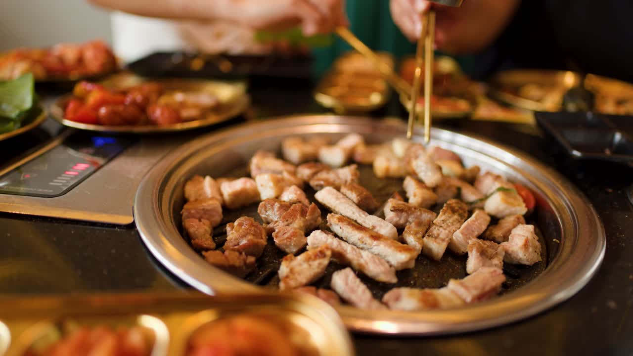 Group uses chopsticks to cook pork on tabletop grill, warm lighting, lively dining atmosphere