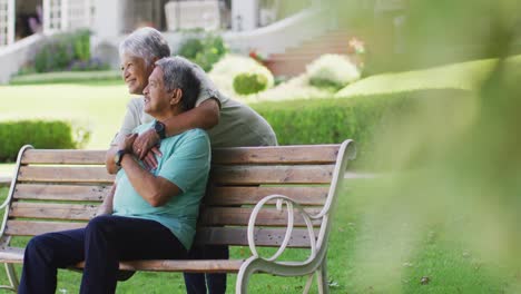 Video-of-happy-biracial-senior-couple-embracing-and-sitting-on-bench-in-garden