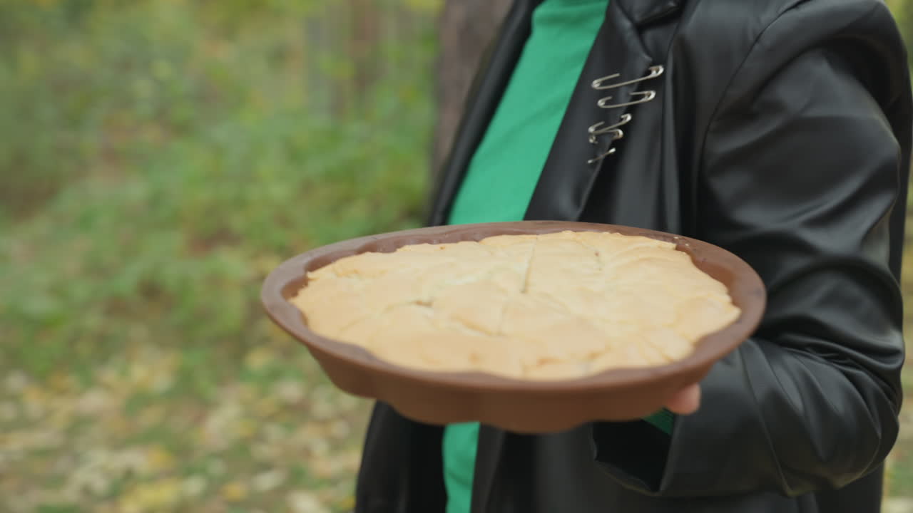 donna con giacca di pelle che offre una torta su un piatto di argilla, primo piano della mano e della crosta su alberi autunnali e foglie cadute, sbirciatina di un maglione verde, atmosfera rustica all'aperto, toni caldi e terrosi, intimo