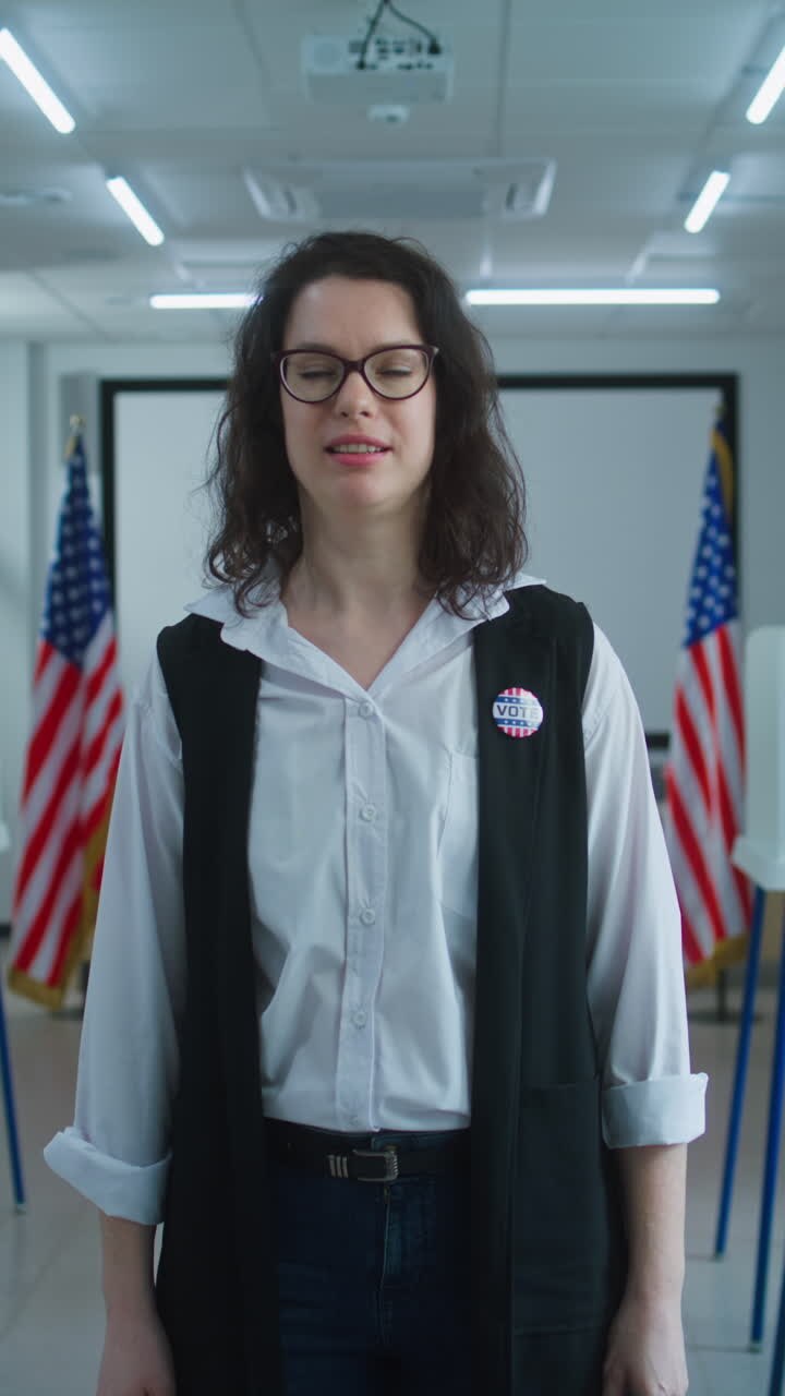 American female voter or polling worker with badge walks and speaks on camera, calls for voting. National Election Day in the United States. Voting booths at polling station. Civic duty and patriotism