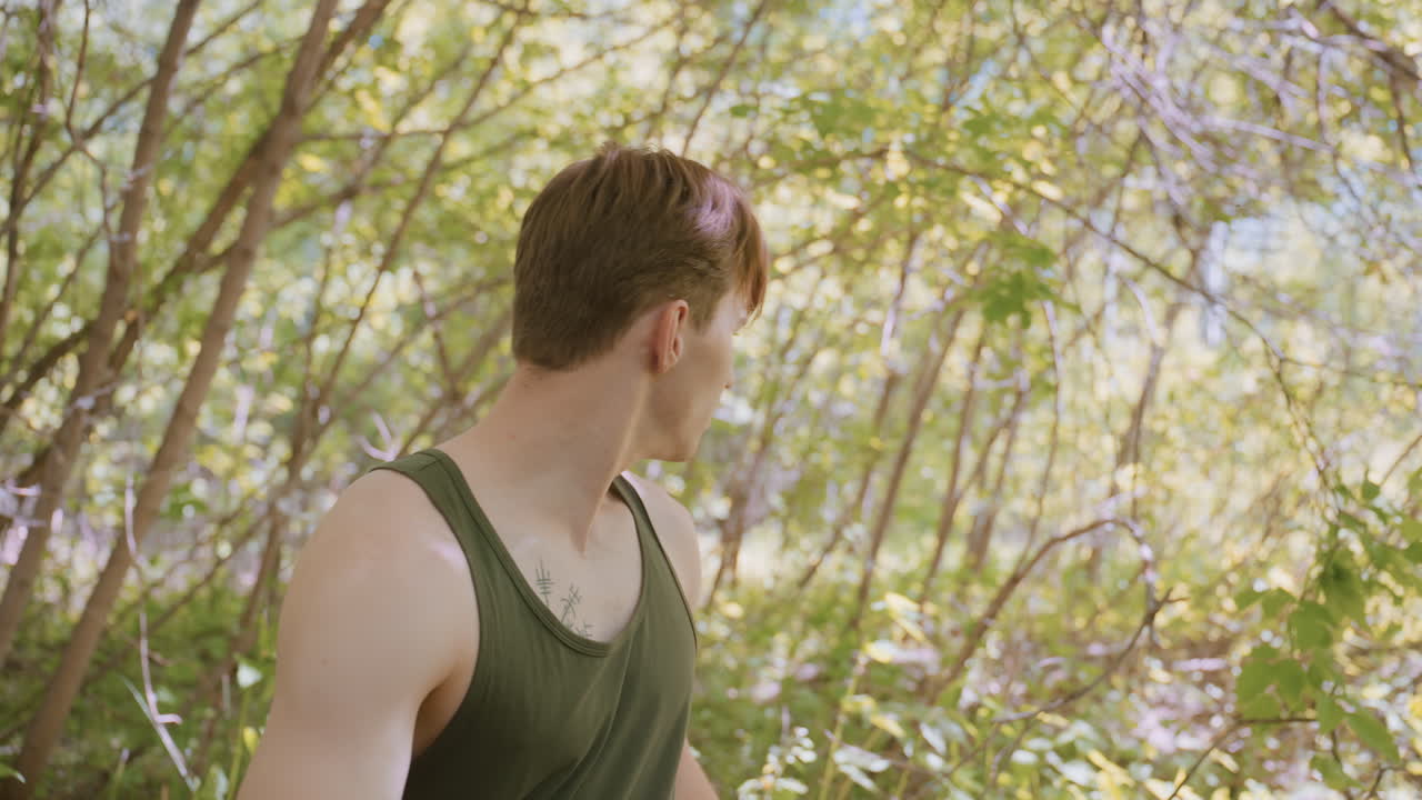 Side view of young man pulling jacket while looking around, showcasing his chest tattoo resembling an arrow, standing in a vibrant forest with natural light filtering through the trees