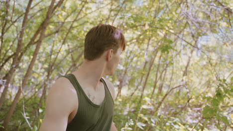 side view of young man pulling jacket while looking around, showcasing his chest tattoo resembling an arrow, standing in a vibrant forest with natural light filtering through the trees
