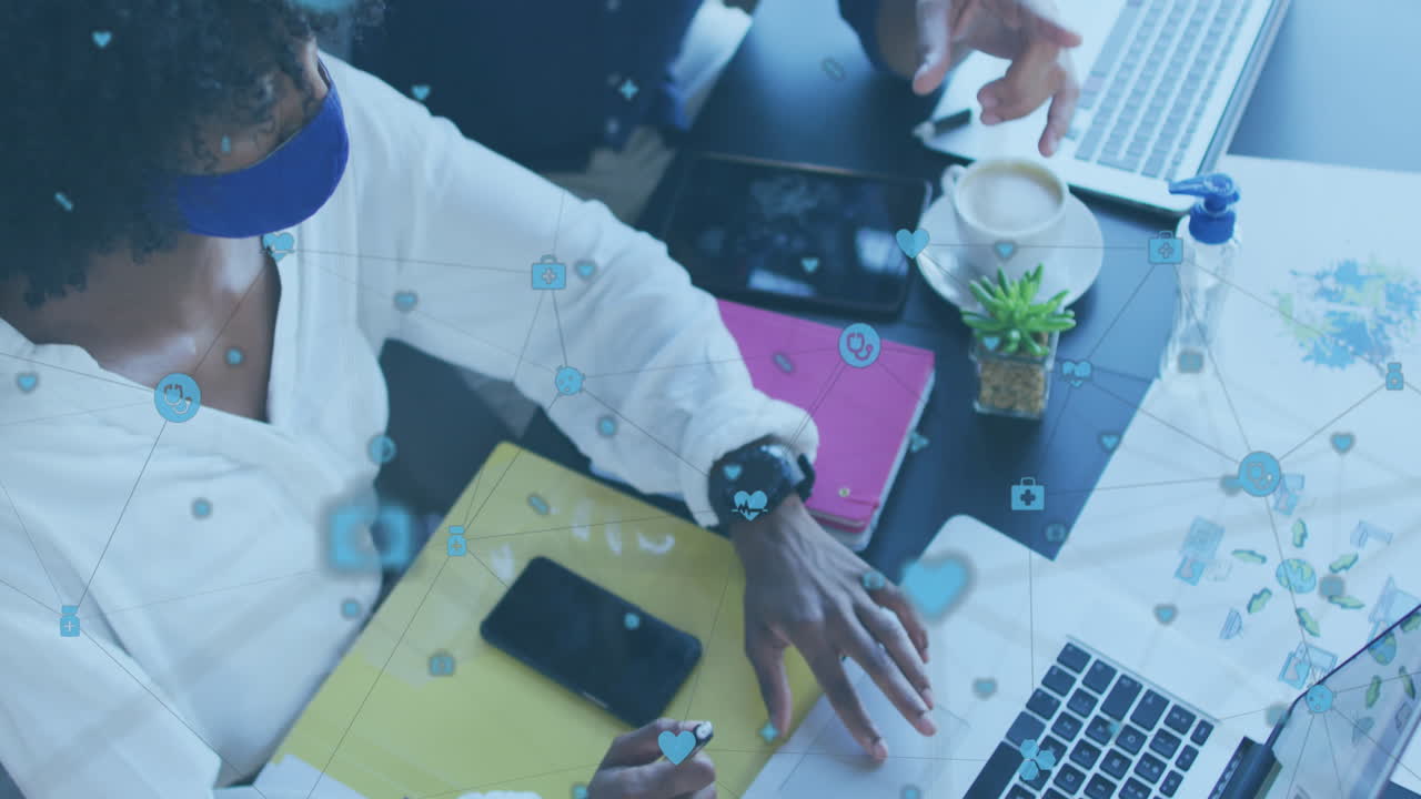 Animation of digital icons over two diverse woman and man wearing face masks discussing at office