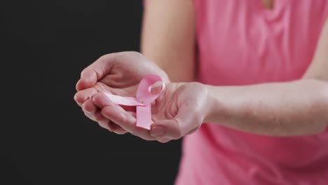 Mid-section-of-woman-holding-a-pink-ribbon-against-black-background