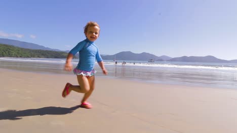 camera orbiting around 4 year old happy young cute girl running freely on the beach with mountains background and calm sea and clear sand