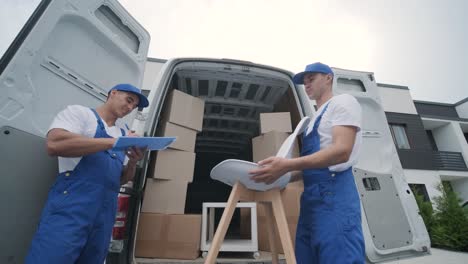 two young workers of removal company unload boxes and furniture from minibus into customer's home