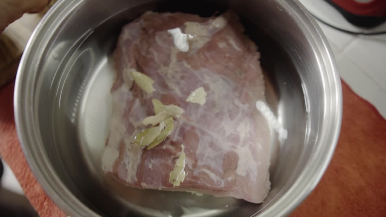 Vista de cima de carne crua colocada em tigela de metal com pedaços de alho sendo adicionados por cima durante o processo de marinada e preparação de alimentos na cozinha.