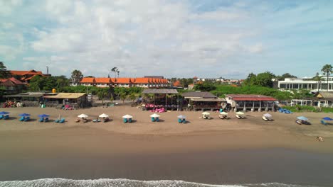 707 beachberm beachfront cafe with people on vacation sun tanning lying on chaise loungers under umbrellas at batu belig beach, bali indonesia - aerial flyover