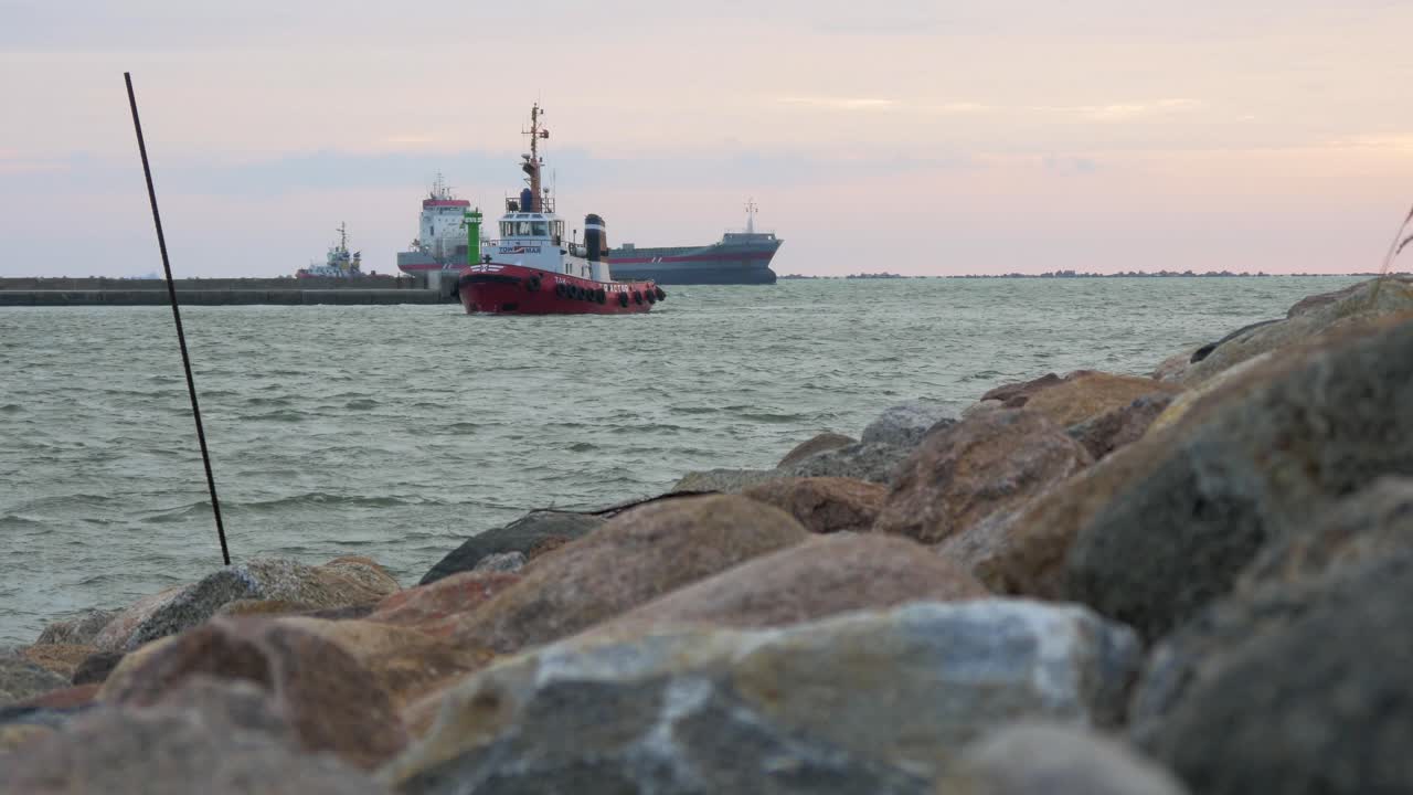 barco piloto de puerto rojo y blanco entrando en el puerto de liepaja en una tranquila y soleada tarde de verano, gran barco de carga gris en el fondo, muelle de piedra en primer plano, olas salpicadas, amplio tiro en ángulo bajo