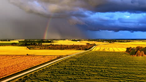 a stunning aerial view captures a beautiful rainbow appearing under dramatic storm clouds, while golden sunlight illuminates the vast agricultural fields of latvia