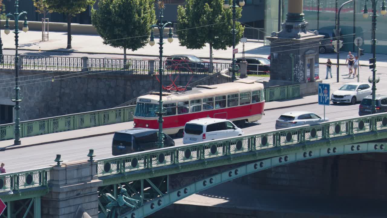 Red tram and vehicles move across ornate city bridge in Prague, bright daylight, steady camera