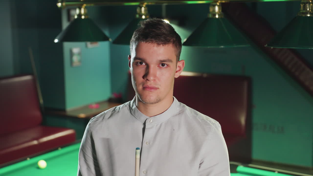 el jugador con cara seria en camisa blanca mantiene la posición enfocada, sosteniendo el palo de la clave en la sala de billar débilmente iluminada. la atmósfera estratégica e intensa destaca la concentración y la preparación para el juego