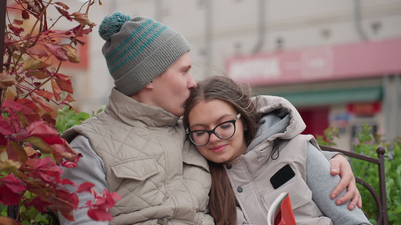 Woman resting peacefully on her love chest with gentle smile as man kisses her head lovingly while seated outdoors on park bench with red autumn leaves and residential buildings in background