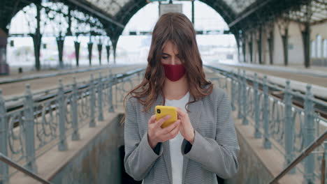 young woman in a face mask using a smartphone at a train station platform