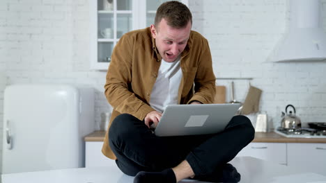 young man looks mad working long hours online. guy sitting on a kitchen table and typing on a wireless laptop. funny fellow playing computer games at home.