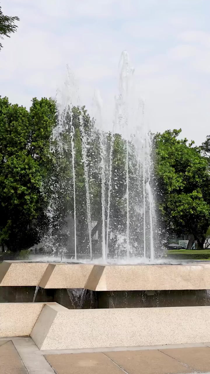 Toma estática de plano medio de una fuente de hormigón escalonada rociando agua contra árboles verdes y el cielo.