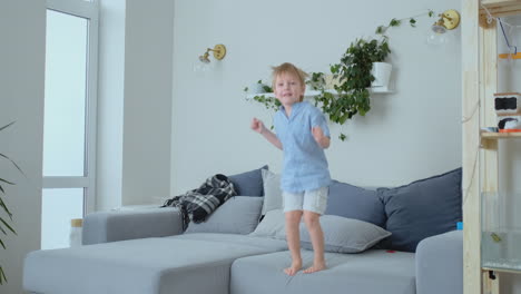 a 4 years old boy with white hair and a blue shirt jumps and smiles on the sofa. fun and baby at home. prank and pampering