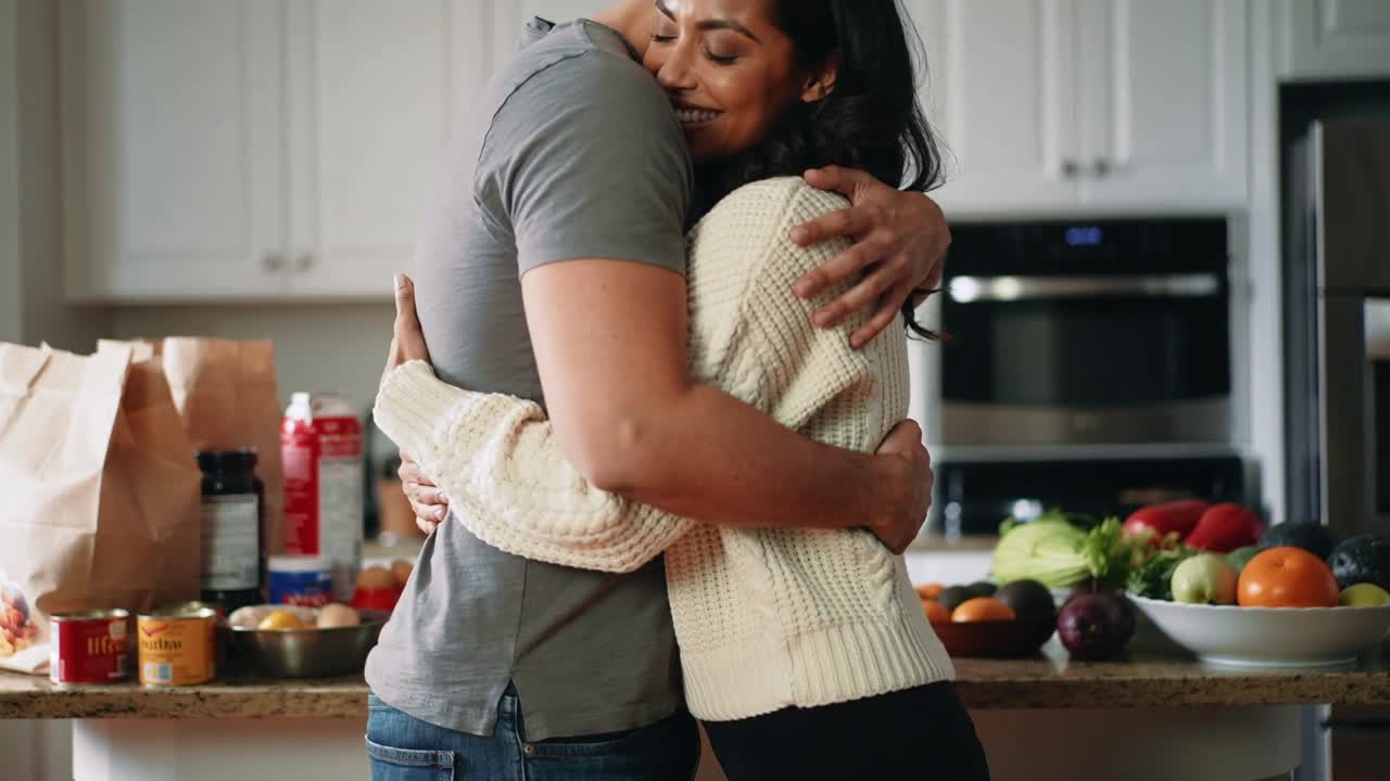 Embracing couple at kitchen island after groceries, wearing grey tee, cream sweater
