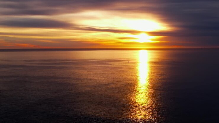 Golden sunrise over the Eastern horizon reflecting off the surface of the tranquil ocean - descending aerial view