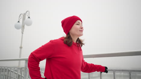 businesswoman exercising outdoors during winter, while stretching her leg back and forth with a warm smile, surrounded by snowy ground, foggy atmosphere, distant river, and light poles