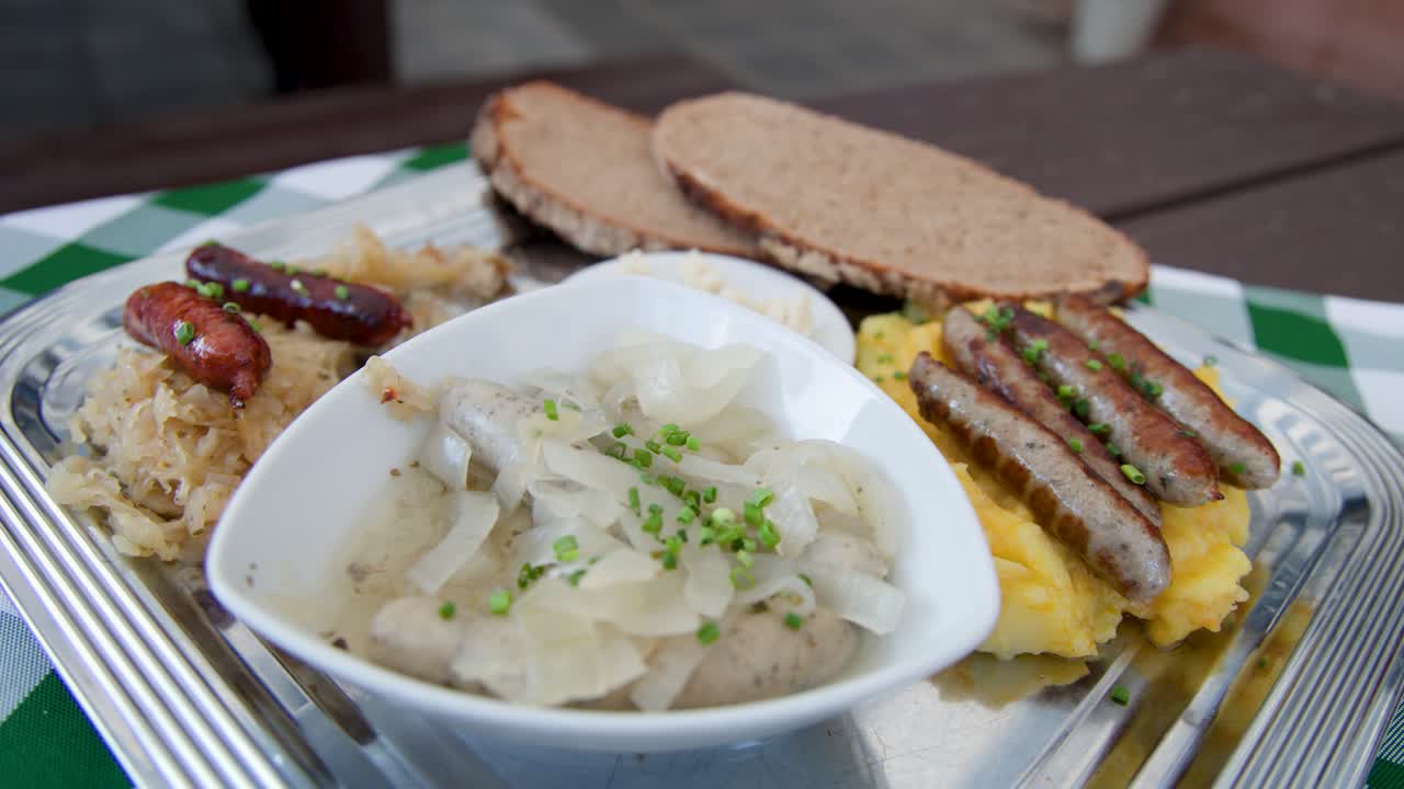 Hand lifts Nuremberg sausage from bowl with onions, surrounded by sauerkraut, bread, potato salad