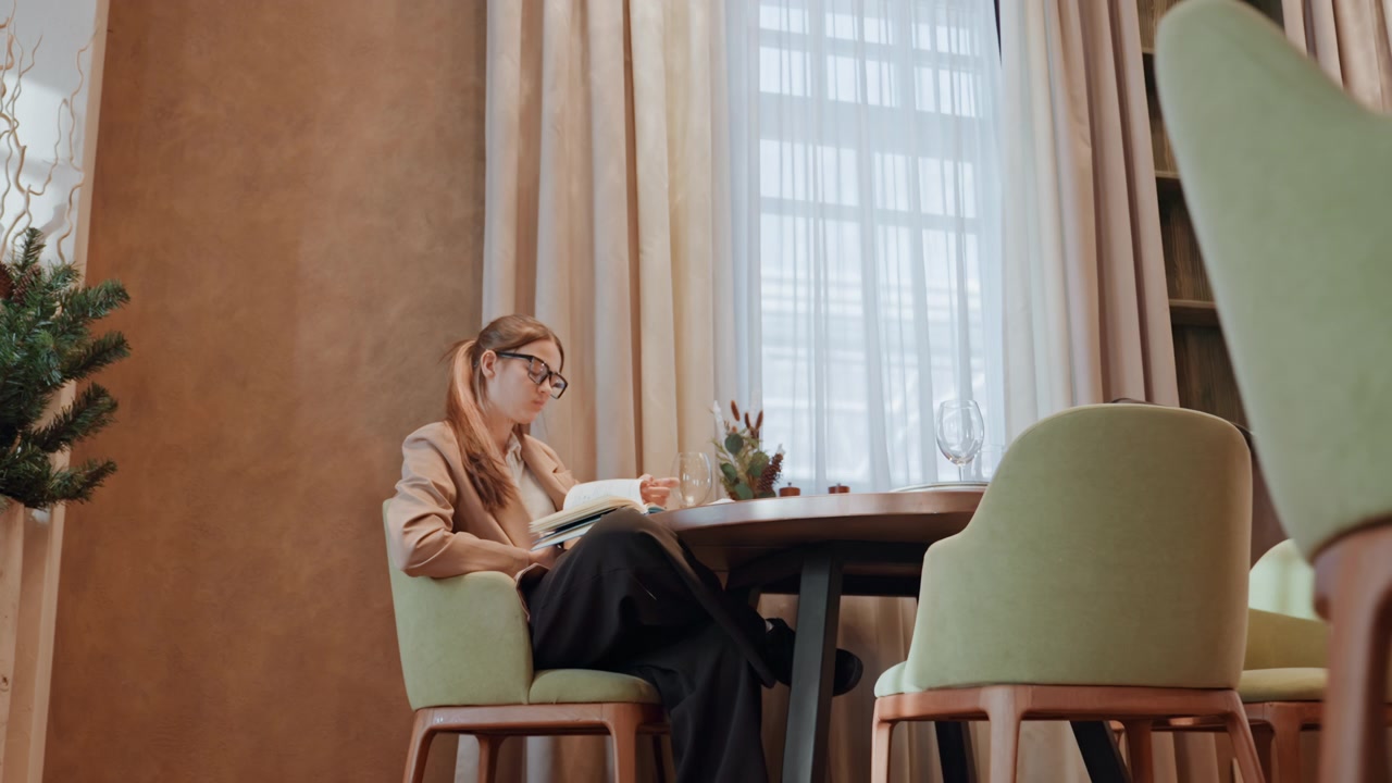 White woman taking notes by window, elegant chandelier and green chairs, planner and sketchbook on table, stylish blazer, focused expression, ambient sunlight through curtains, boutique cafe interior